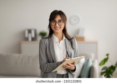 Happy professional middle eastern female psychologist writing in clipboard, looking and smiling at camera, working in modern office. Psychotherapy services, mental health professional concept Foto stock