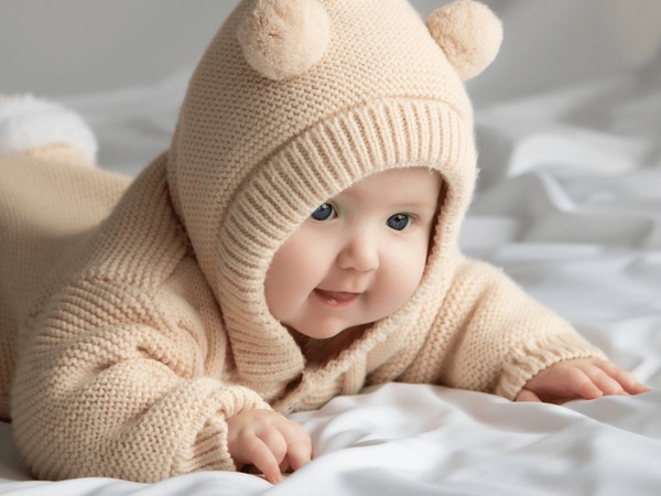 Just beautiful. Cute smiling baby. Cute 3 month old Baby girl infant on a bed on her belly with head up looking with her big eyes. Warm, fluffy biege clothes. Closeup. Three months old baby Stock Photo